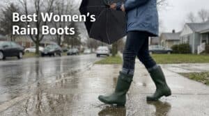 Woman walking in rain with dark green waterproof rain boots on a wet street, holding an umbrella, in a residential neighborhood.