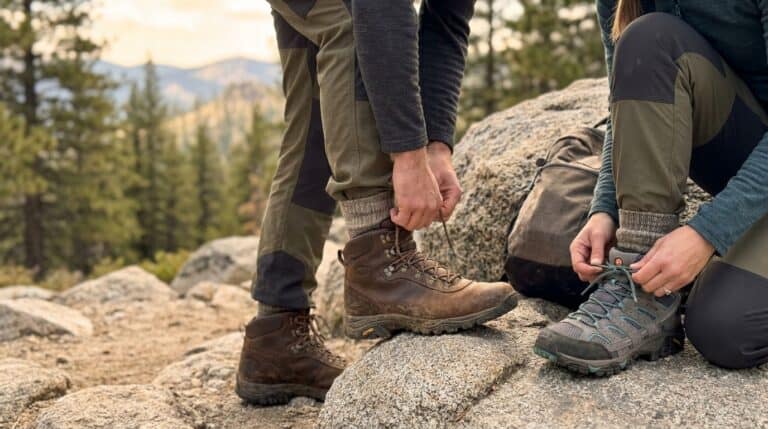 Two hikers tying their sturdy hiking boots on a rocky trail surrounded by trees and mountains, emphasizing durability and comfort for outdoor adventures.