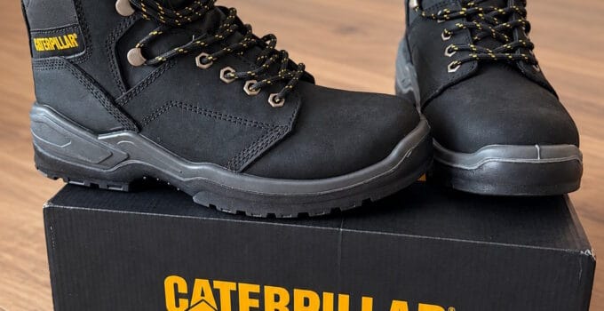 Pair of black Caterpillar CAT steel toe work boots with yellow accents, placed on a black Caterpillar box on a wooden floor.
