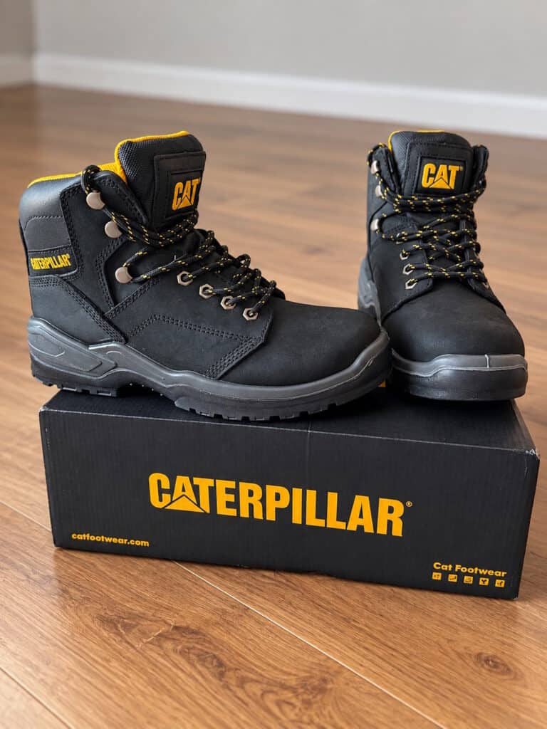 Pair of black Caterpillar CAT steel toe work boots with yellow accents, placed on a black Caterpillar box on a wooden floor.