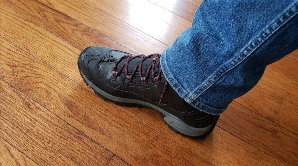 Close-up of a men's black hiking boot with red laces worn with blue jeans on a wooden floor.