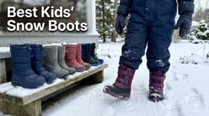 A child wearing purple snow boots standing in snow with a display of various kids' winter boots on a bench nearby.