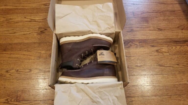 Brown leather hiking boot in open box with tissue paper on wooden floor.