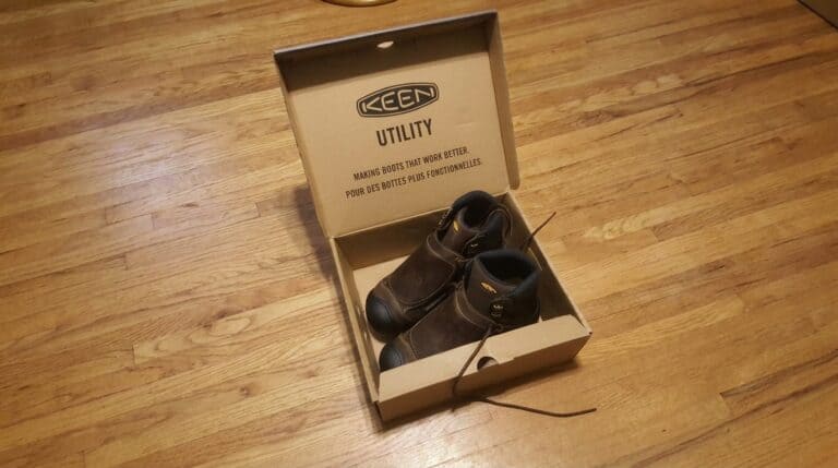 Pair of KEEN utility work boots inside a cardboard box on a wooden floor, showcasing durable footwear for work environments.