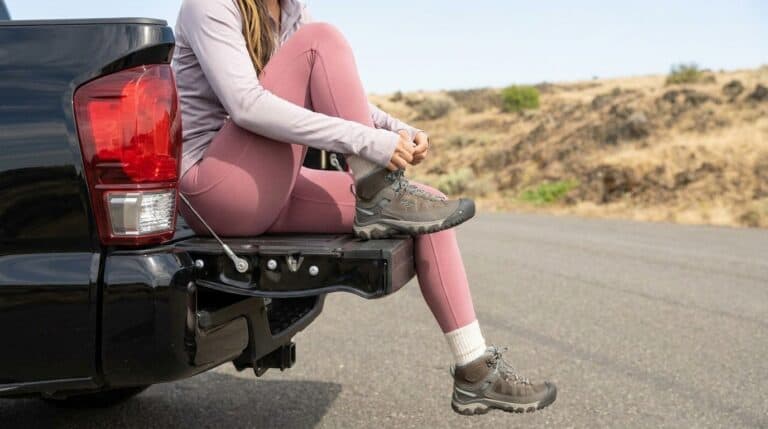 Woman in pink leggings and hiking boots sitting on the tailgate of a black pickup truck on a rural road during daytime.