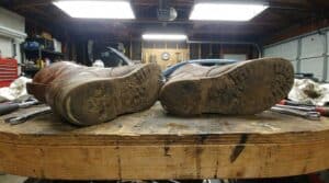 Close-up of the soles of worn work boots showing significant tread wear and dirt, indicating extensive use in demanding environments.